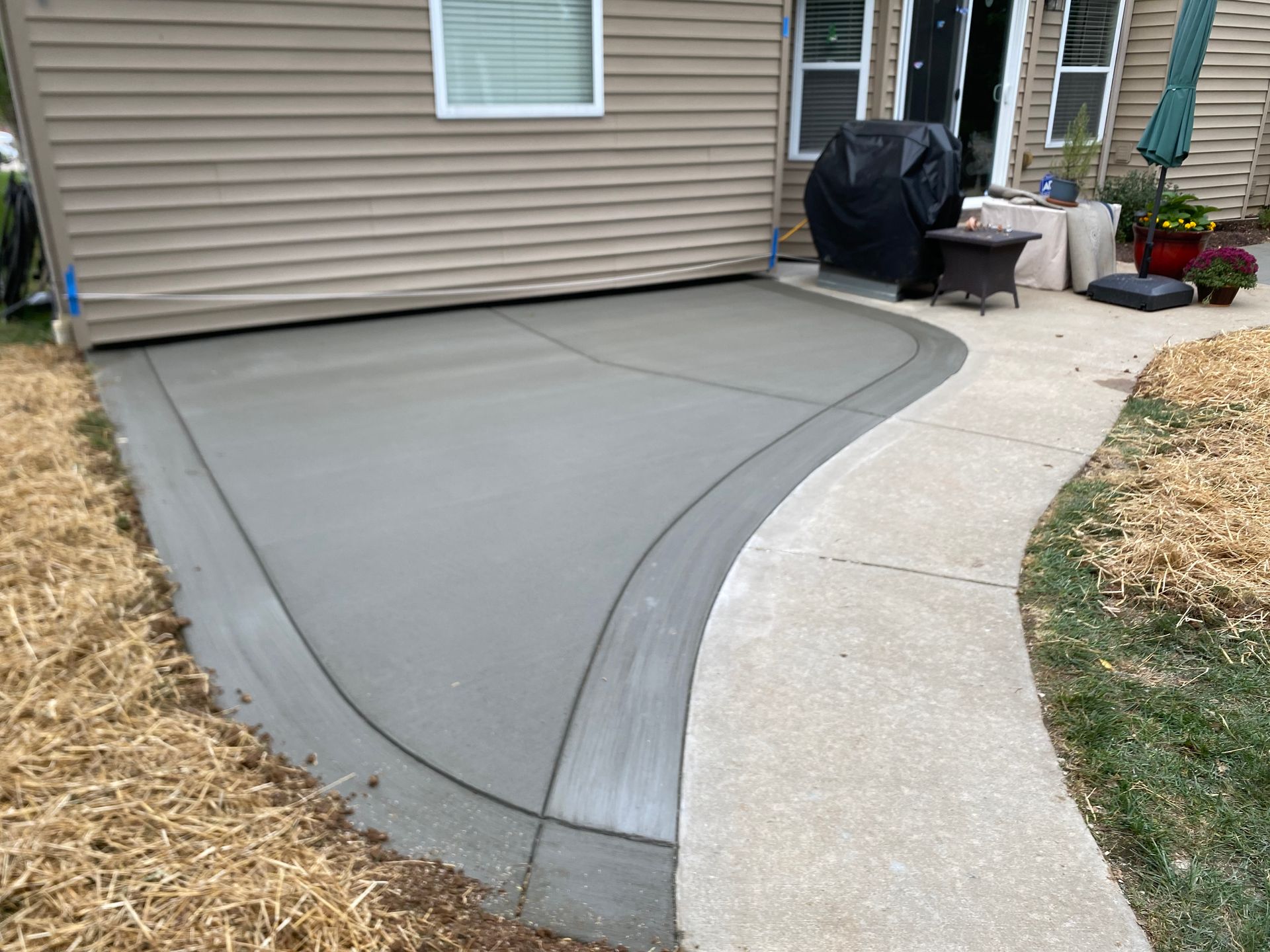 Curved concrete patio beside a house, bordered by a light-colored walkway and mulch beds.