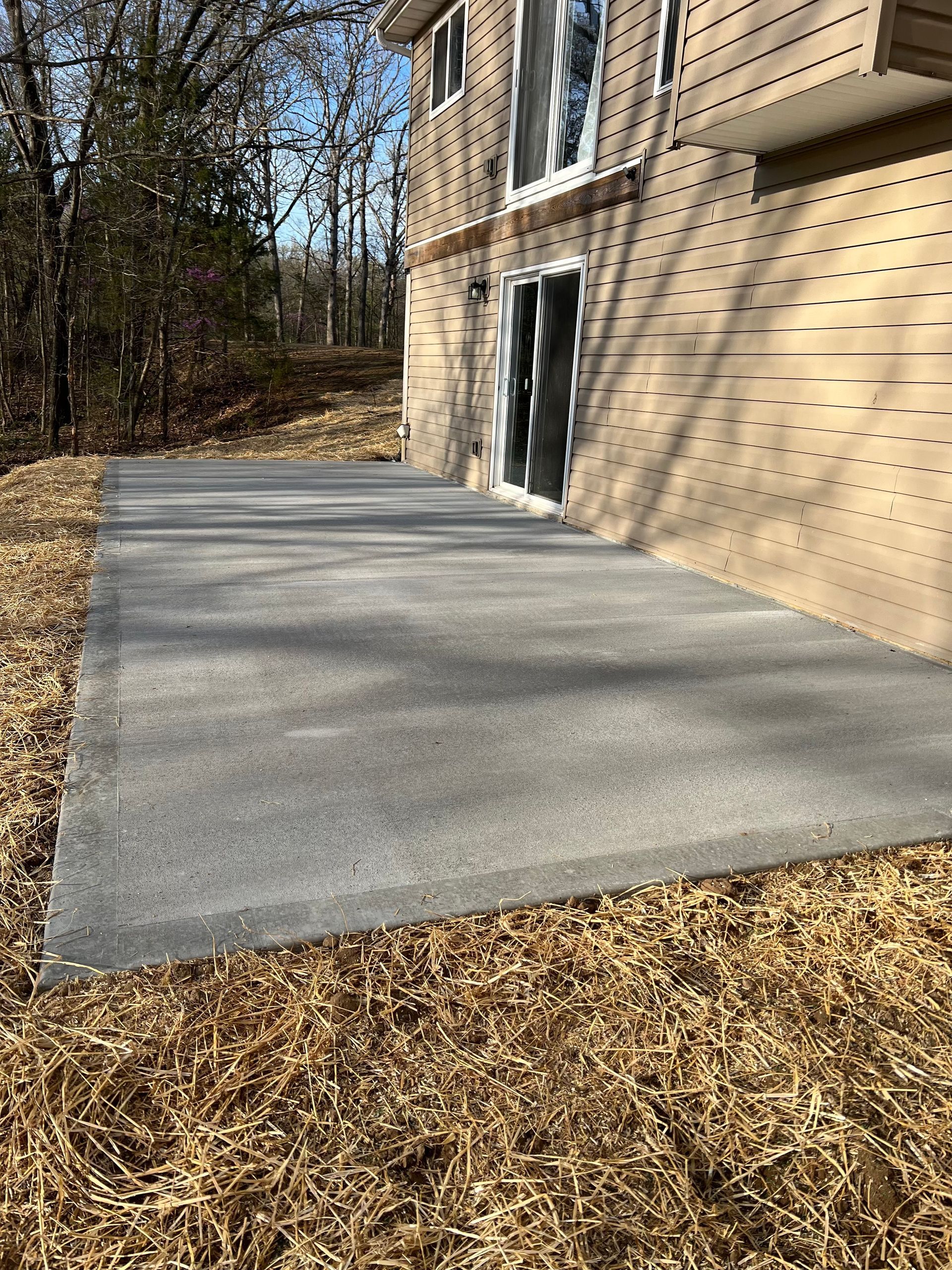Fresh concrete patio beside a brick house with a glass door, bordered by mulch and leafless trees