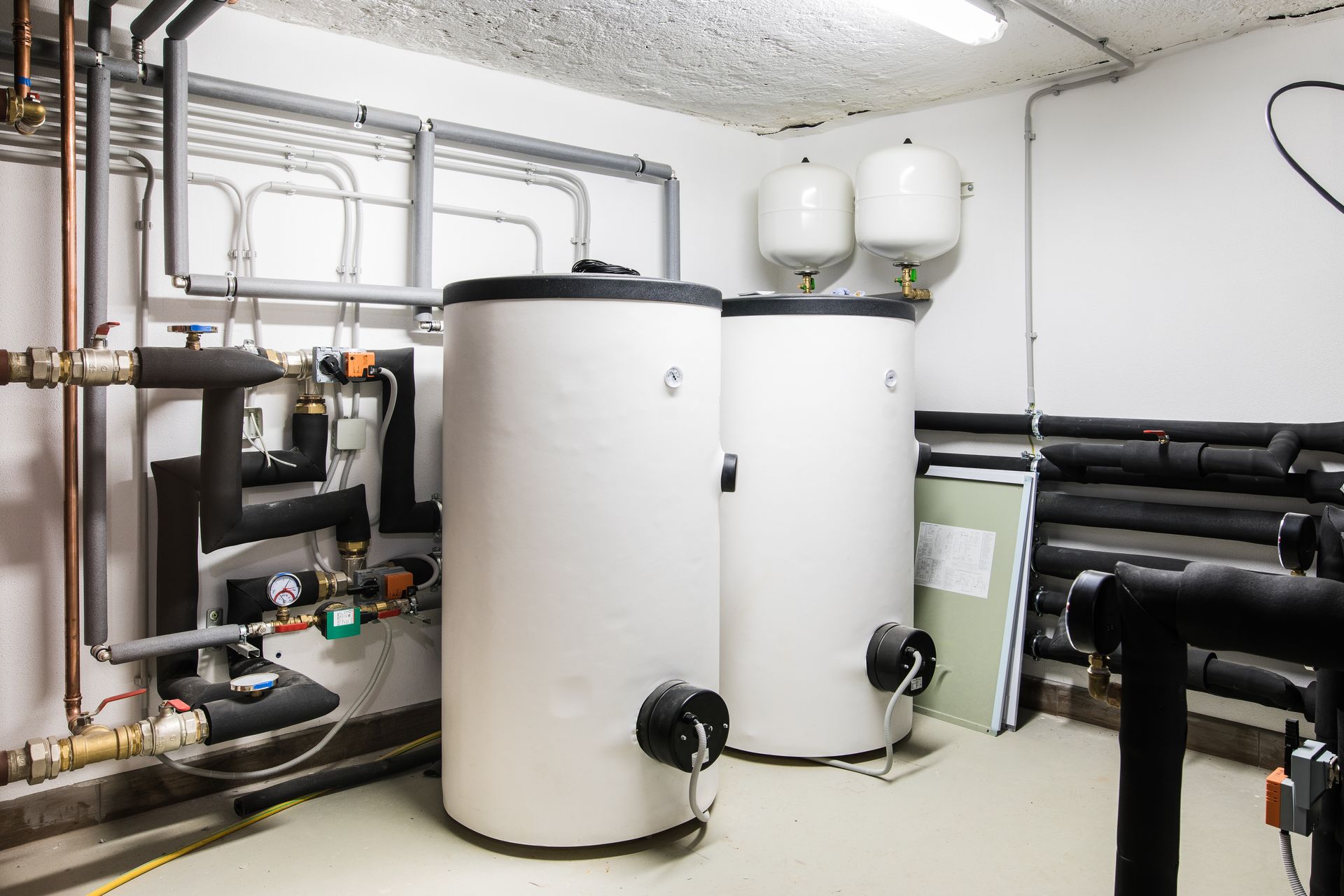 Boiler room with white storage tanks, pipes, valves, and wall-mounted tanks in a utility space