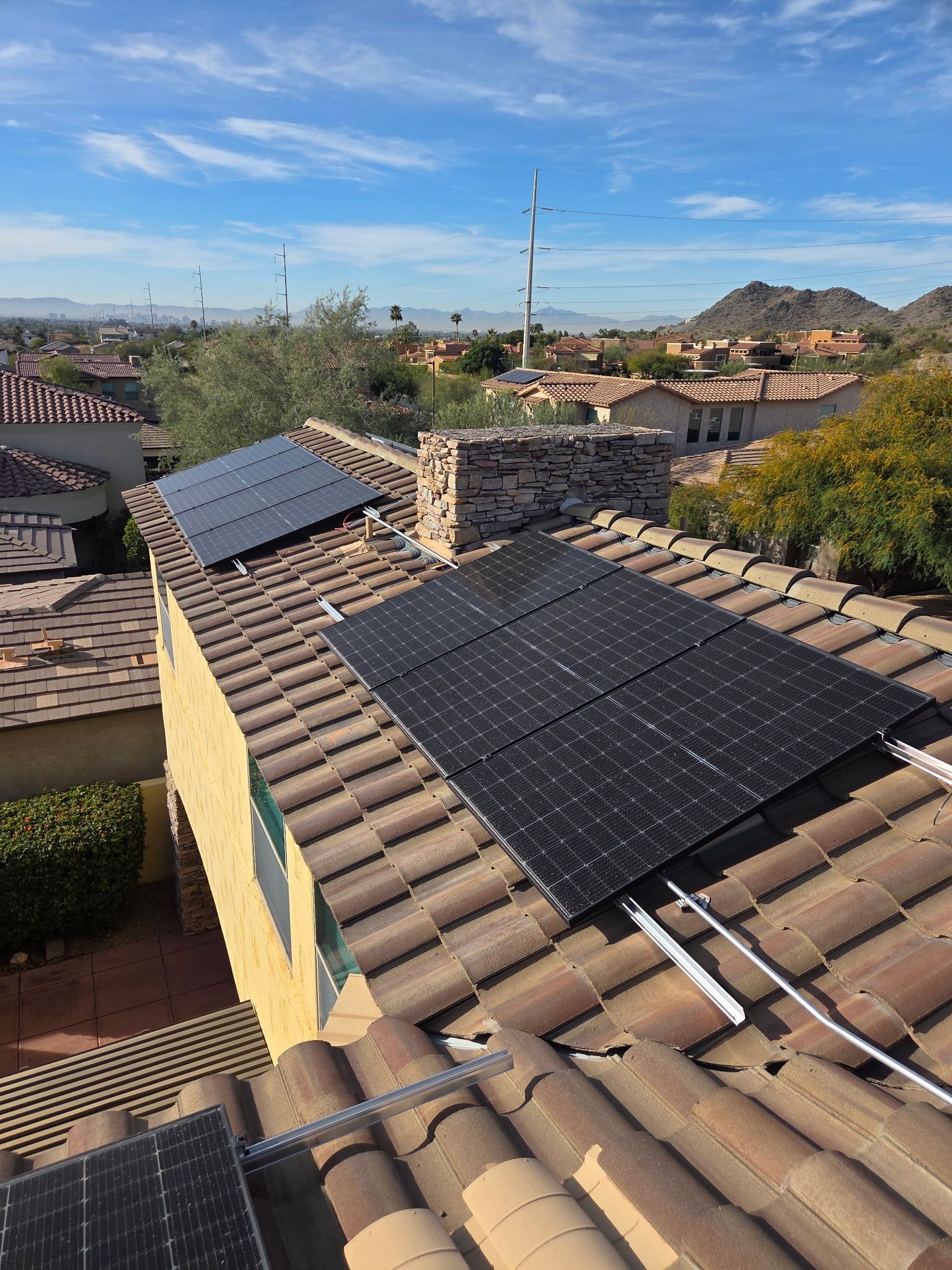 Solar panels installed on a brown tiled residential roof under a sunny sky with distant desert hills in the background.