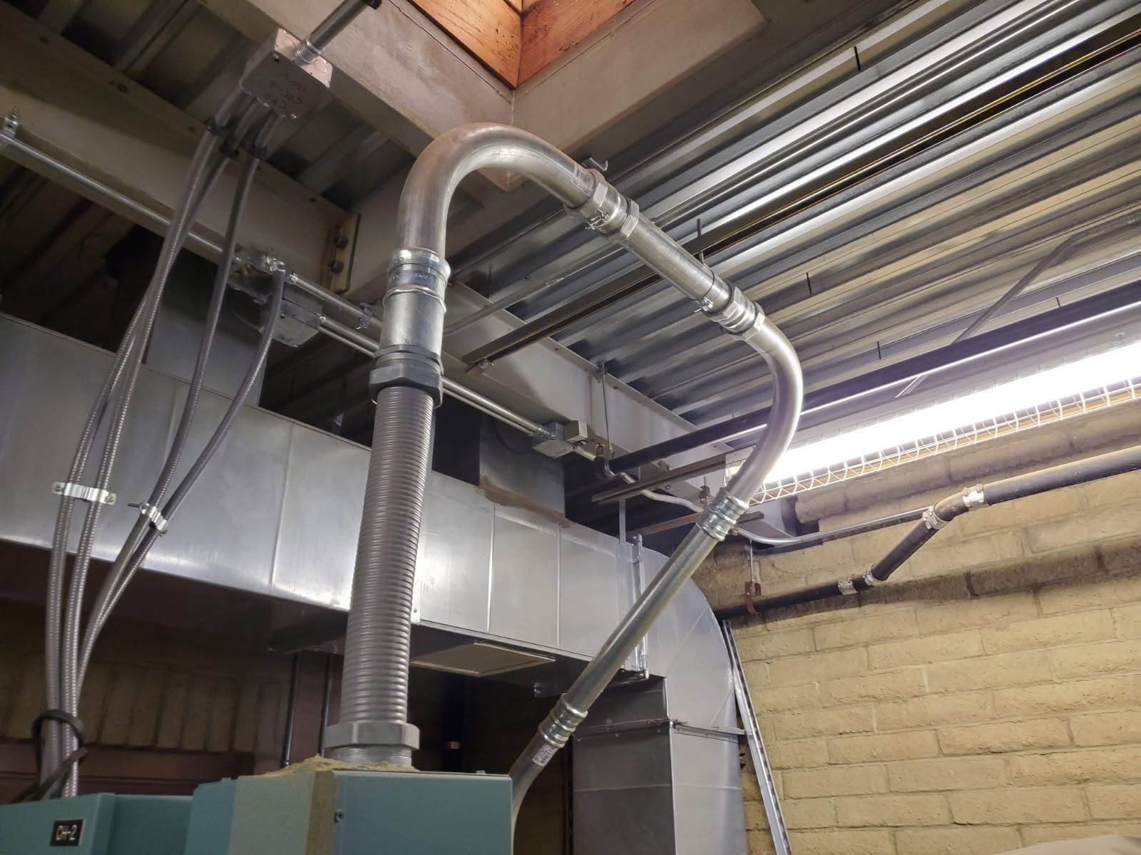 A silver metallic conduit pipe loops upward from industrial machinery to join with overhead electrical infrastructure.