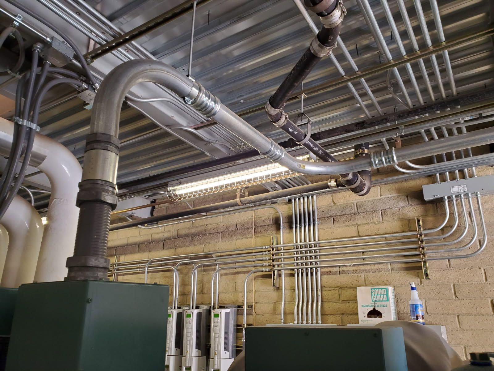 A view of industrial piping and electrical conduits running across a ceiling and brick wall in a utility room.