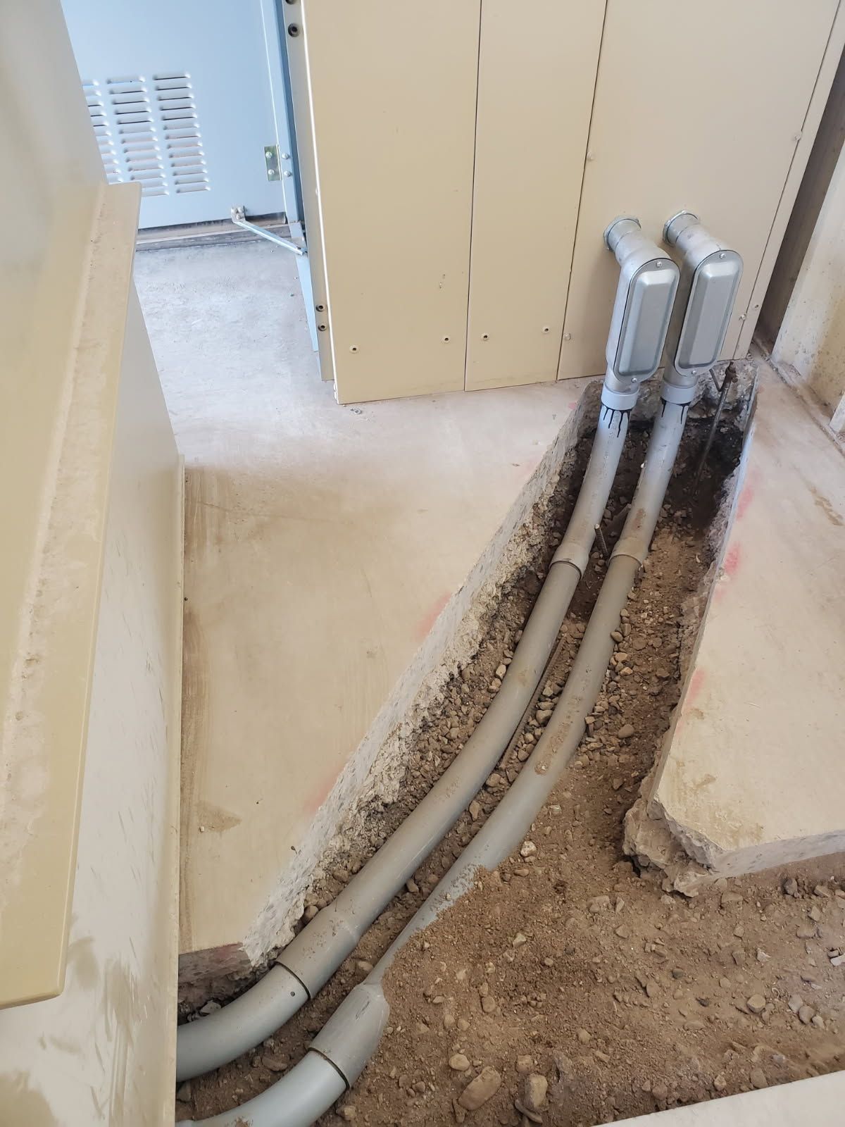 Two grey electrical conduits emerge from a dirt-filled floor trench and connect to a wall-mounted panel.