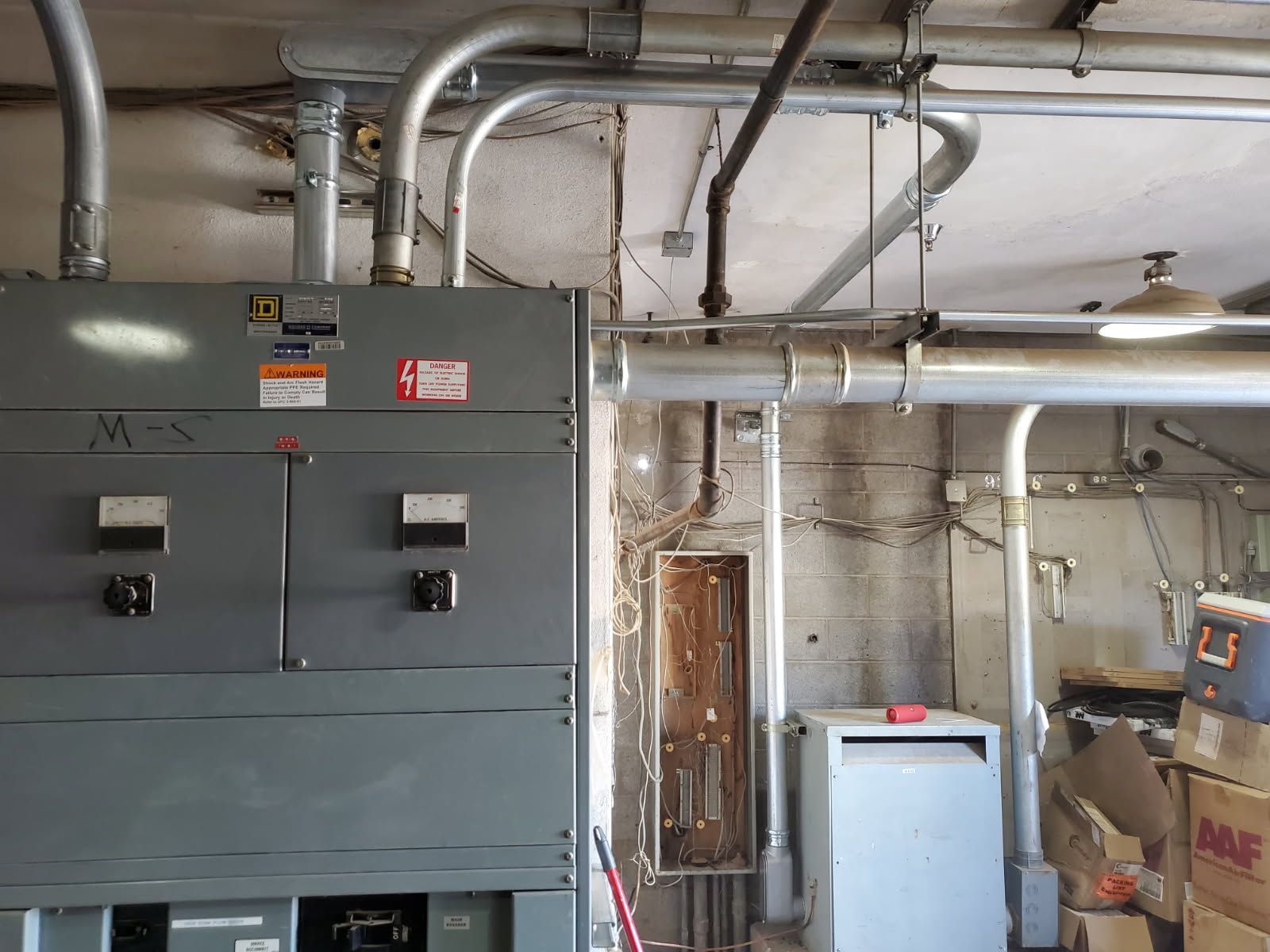 A large grey electrical control panel with conduit pipes in an unfinished basement with equipment and boxes.