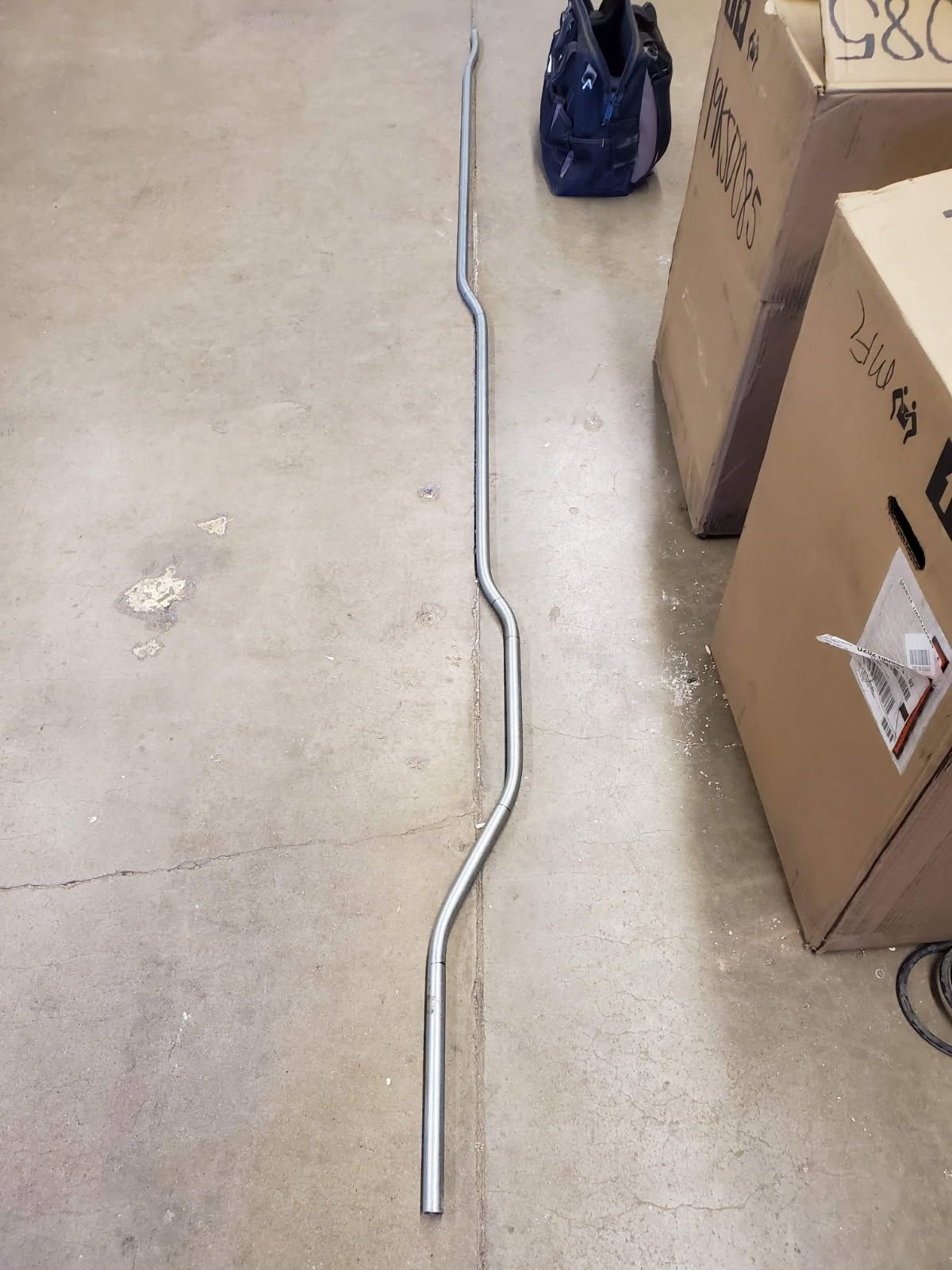 A long, silver, wavy barbell bar lying on a concrete floor next to cardboard boxes and a blue bag.