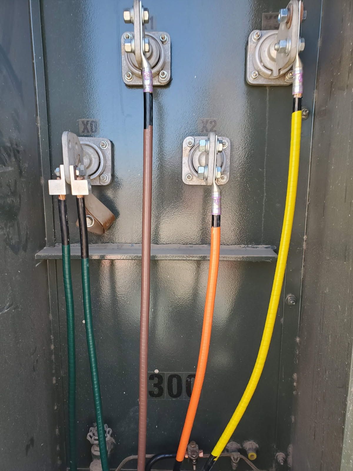 Electrical transformer terminals with green, brown, orange, and yellow cables connected to metal lugs on a dark gray panel.