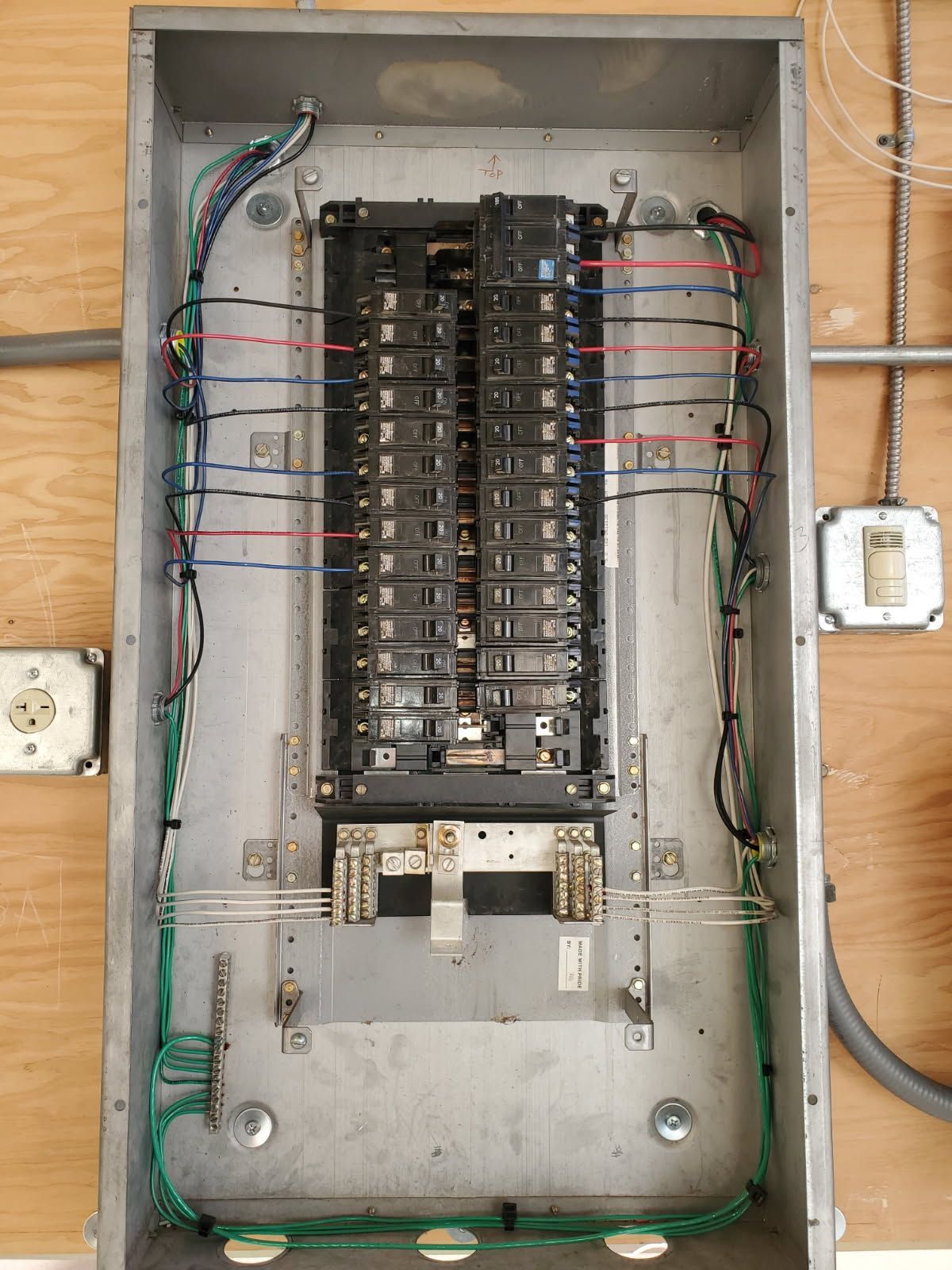 Open electrical service panel with circuit breakers and multicolored wiring installed on a plywood wall.