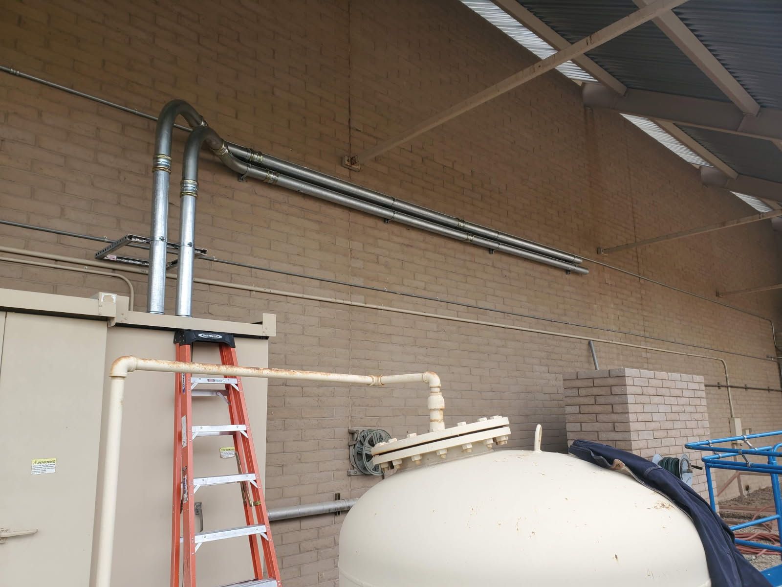Two electrical conduits run along a beige brick wall, extending from a utility cabinet topped by a red ladder.