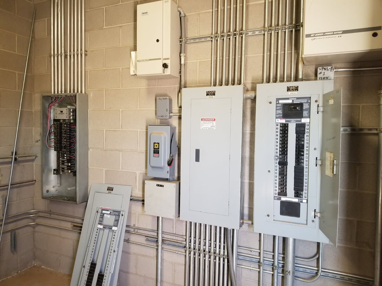 Electrical panels and conduits mounted on a cinder block wall, with one panel cover removed and leaning against the wall.