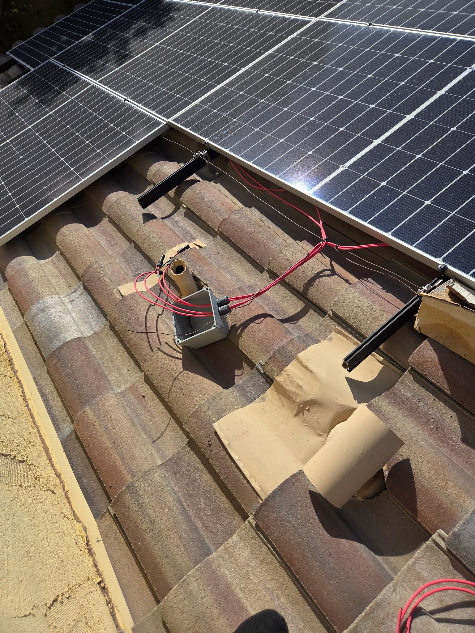 A junction box with red and black electrical wires sits on a brown tiled roof beneath installed solar panels.
