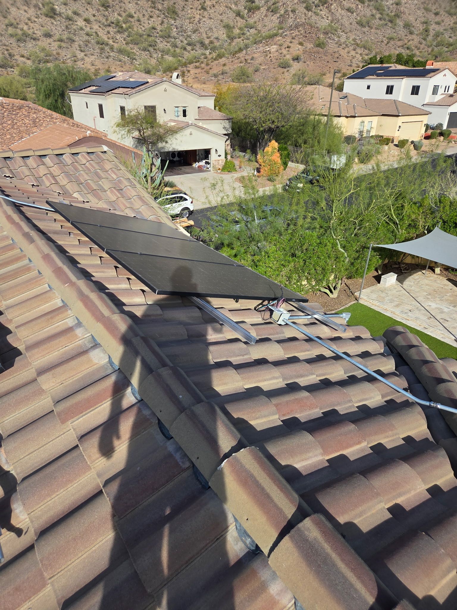 A person stands on a brown tile roof during solar panel installation, with houses and a hillside in the background.