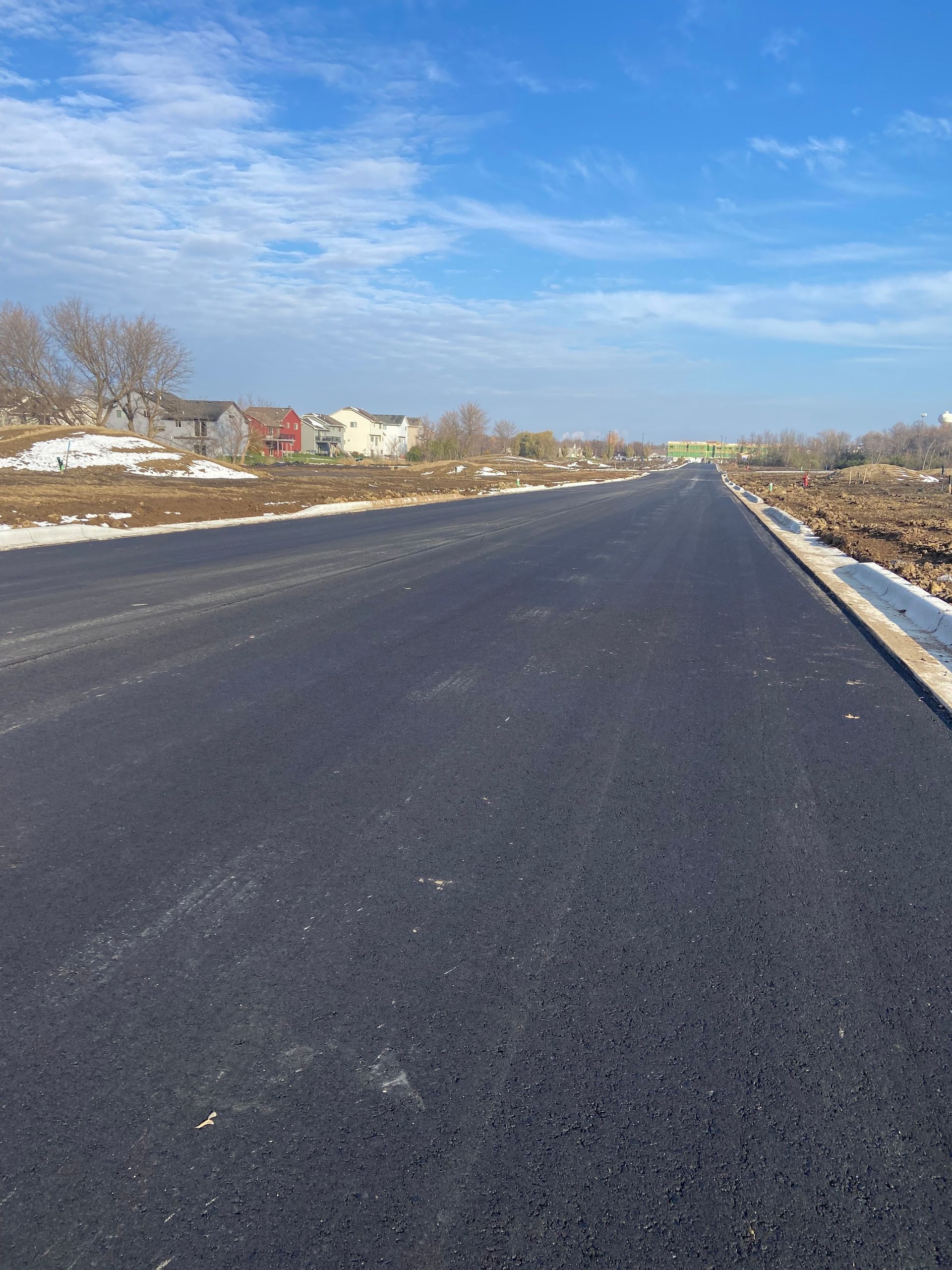 Newly paved asphalt road stretches toward a residential area under a bright blue sky.
