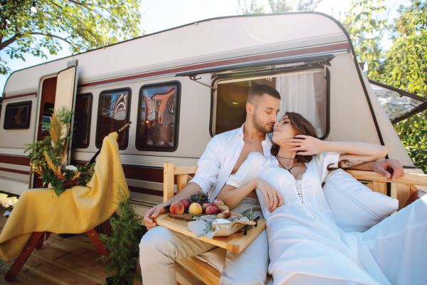Couple relaxing outside a white camper van, sharing a kiss on a picnic blanket with fruits and wine