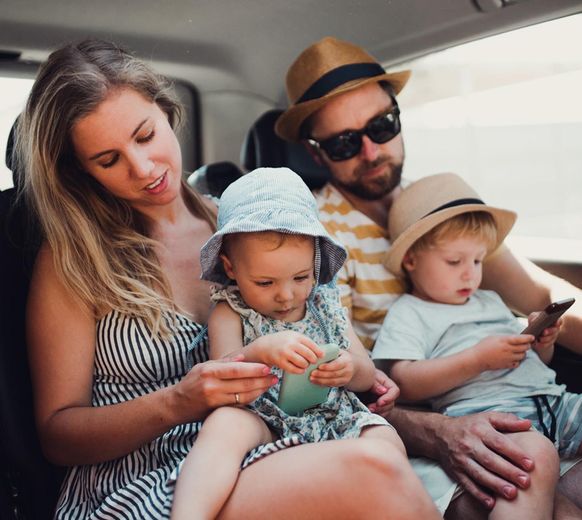 Family sitting together in a car, adults and children wearing hats and looking at a phone