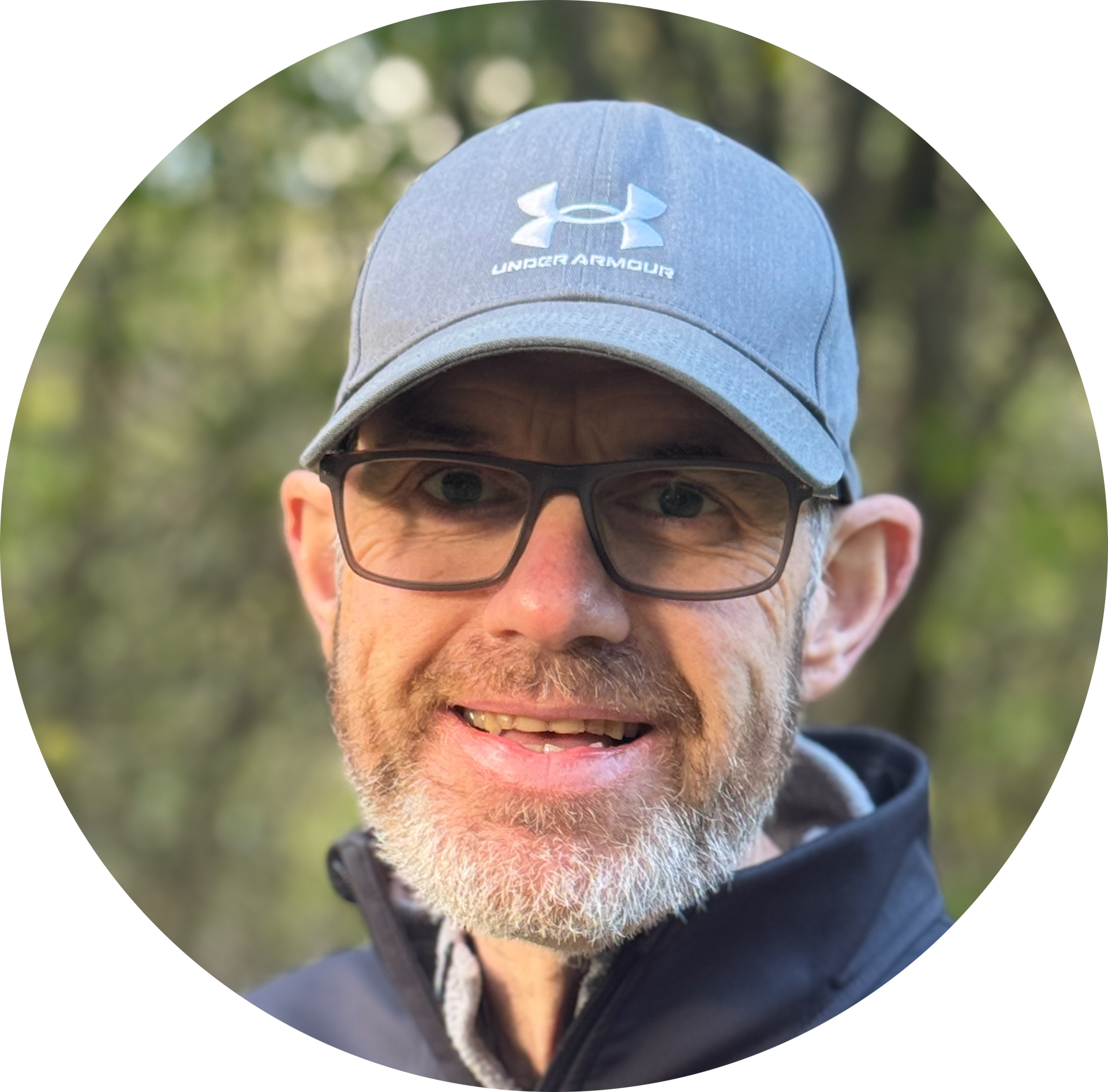 Man wearing glasses and a gray Under Armour cap, smiling outdoors.