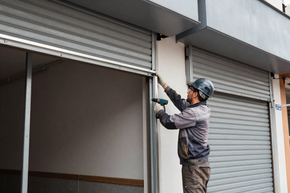 Un trabajador utiliza un taladro para instalar una persiana metálica en la fachada de un local comercial