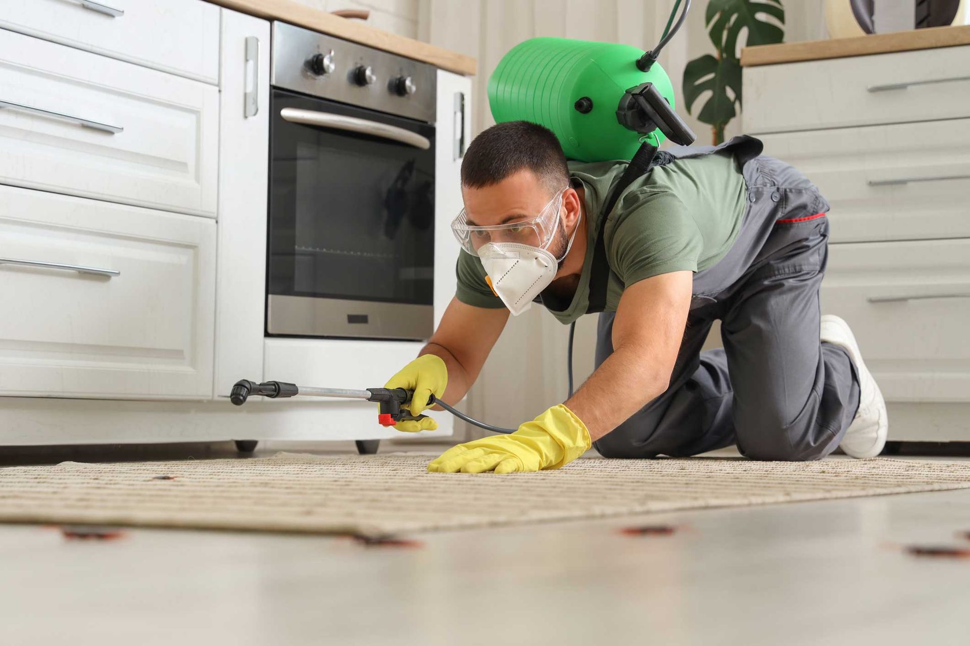 Pest control worker spraying treatment on floor in a kitchen while wearing protective gear.