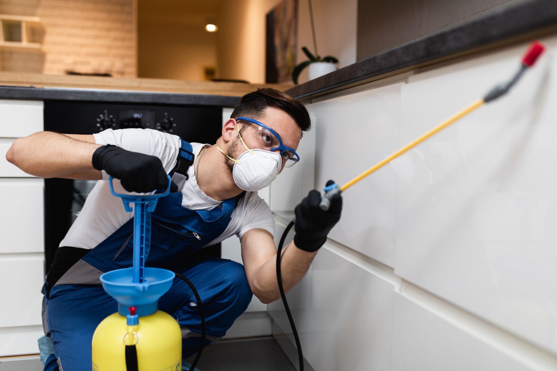 An exterminator in protective workwear is spraying pesticide in the kitchen.