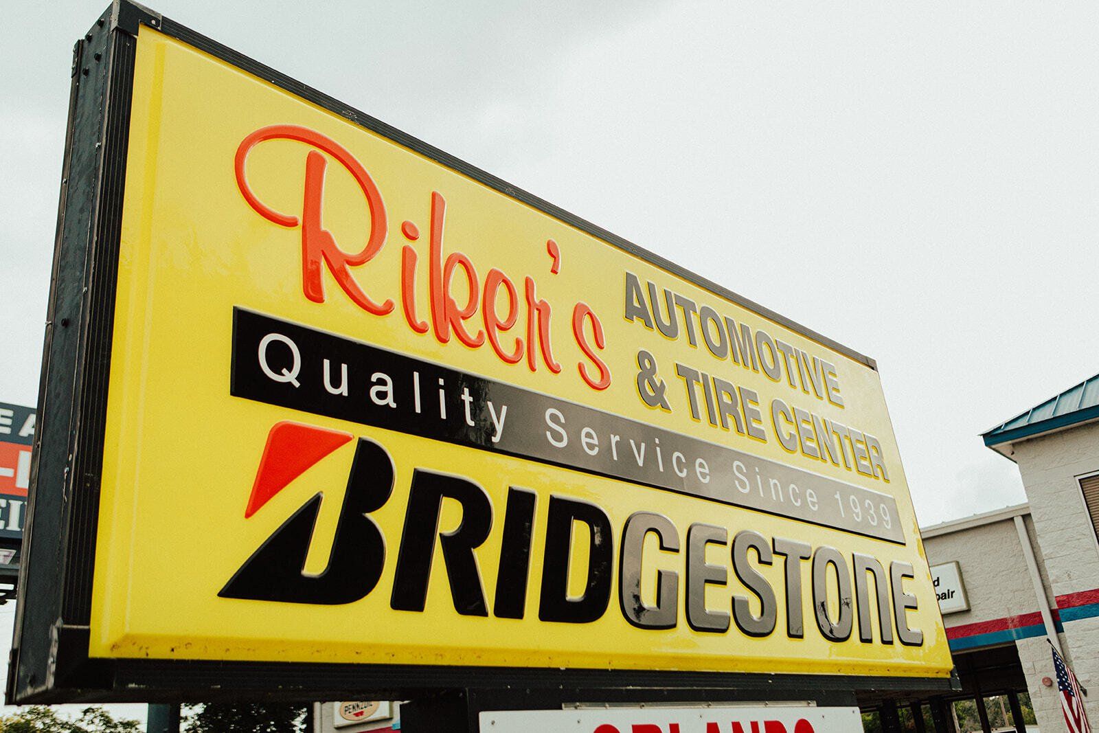Sign for Riker's Automotive & Tire Center, a Bridgestone dealer. Yellow sign with red and black lettering.