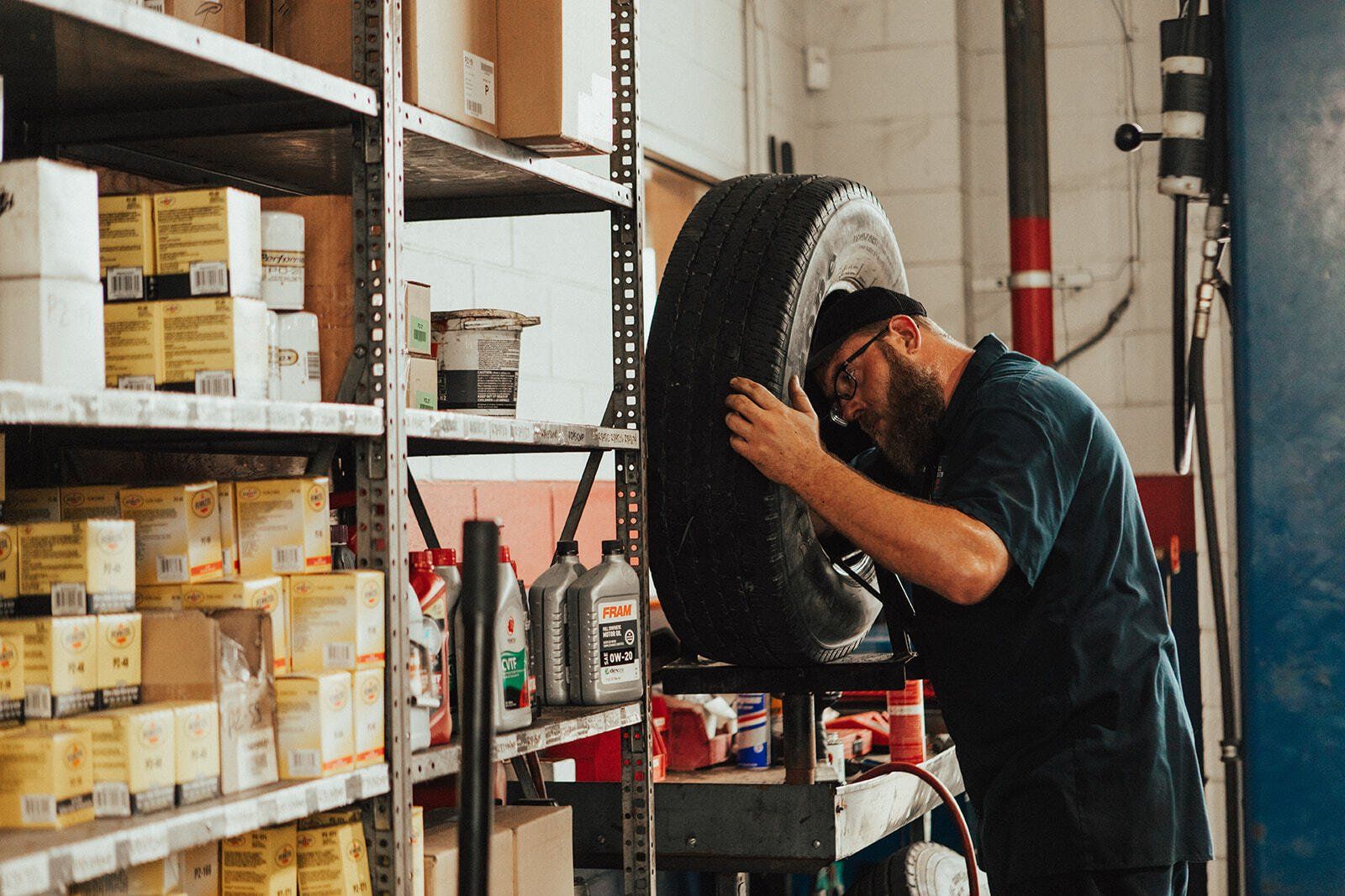 Mechanic inspecting a tire in a garage, surrounded by shelves of parts and fluids.