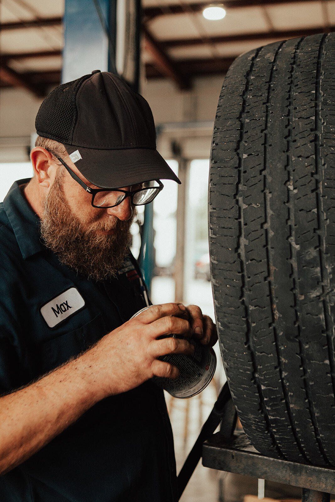 Mechanic examines tire, wearing glasses and hat, in a garage setting.