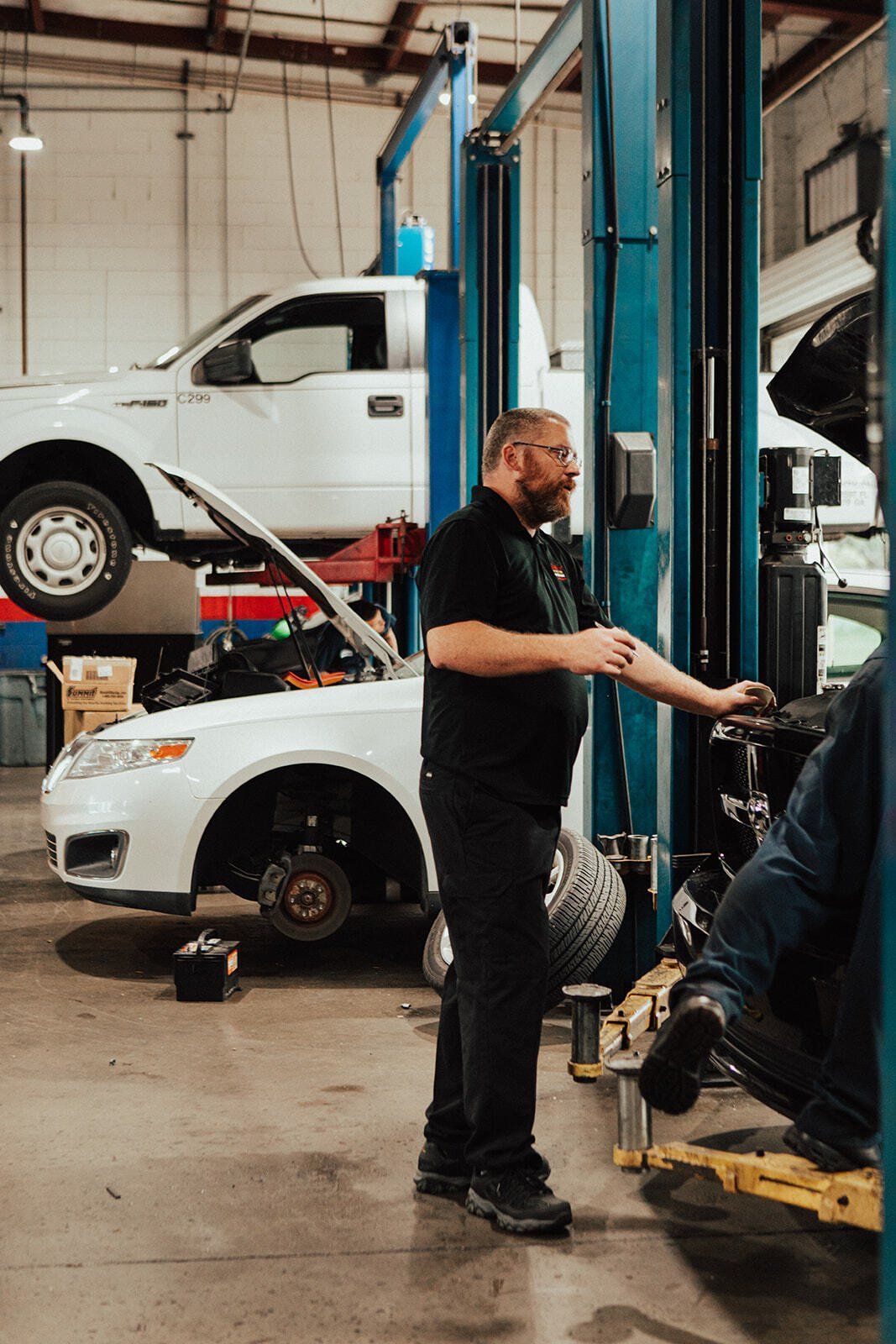 Mechanic in auto shop, standing near a car on a lift; another car on a lift in the background.