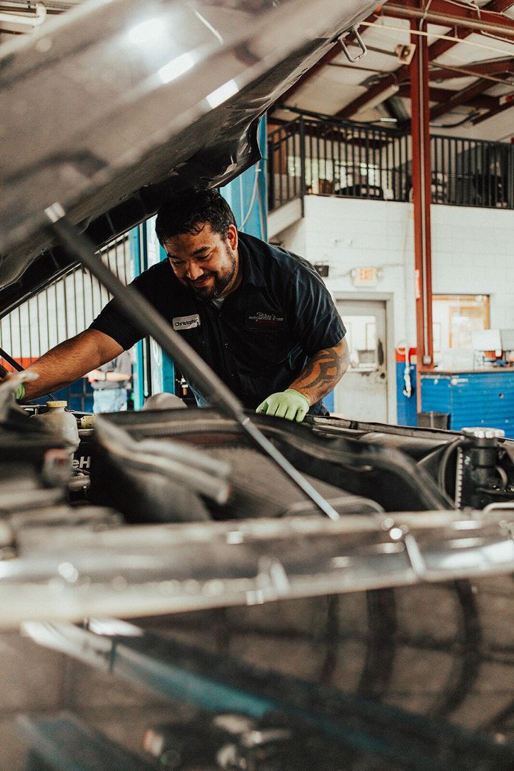 Mechanic working under the hood of a car in a garage, wearing gloves and smiling.