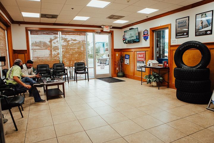 Waiting room of a tire shop with customers. Black chairs, coffee table, stacked tires, TV.