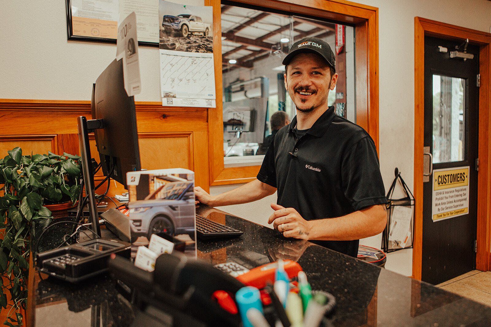 Man at a reception desk smiles, in a car service center. He wears a black shirt and cap.