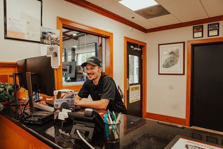 Man smiles behind a reception desk, interior, with a view into a workshop.