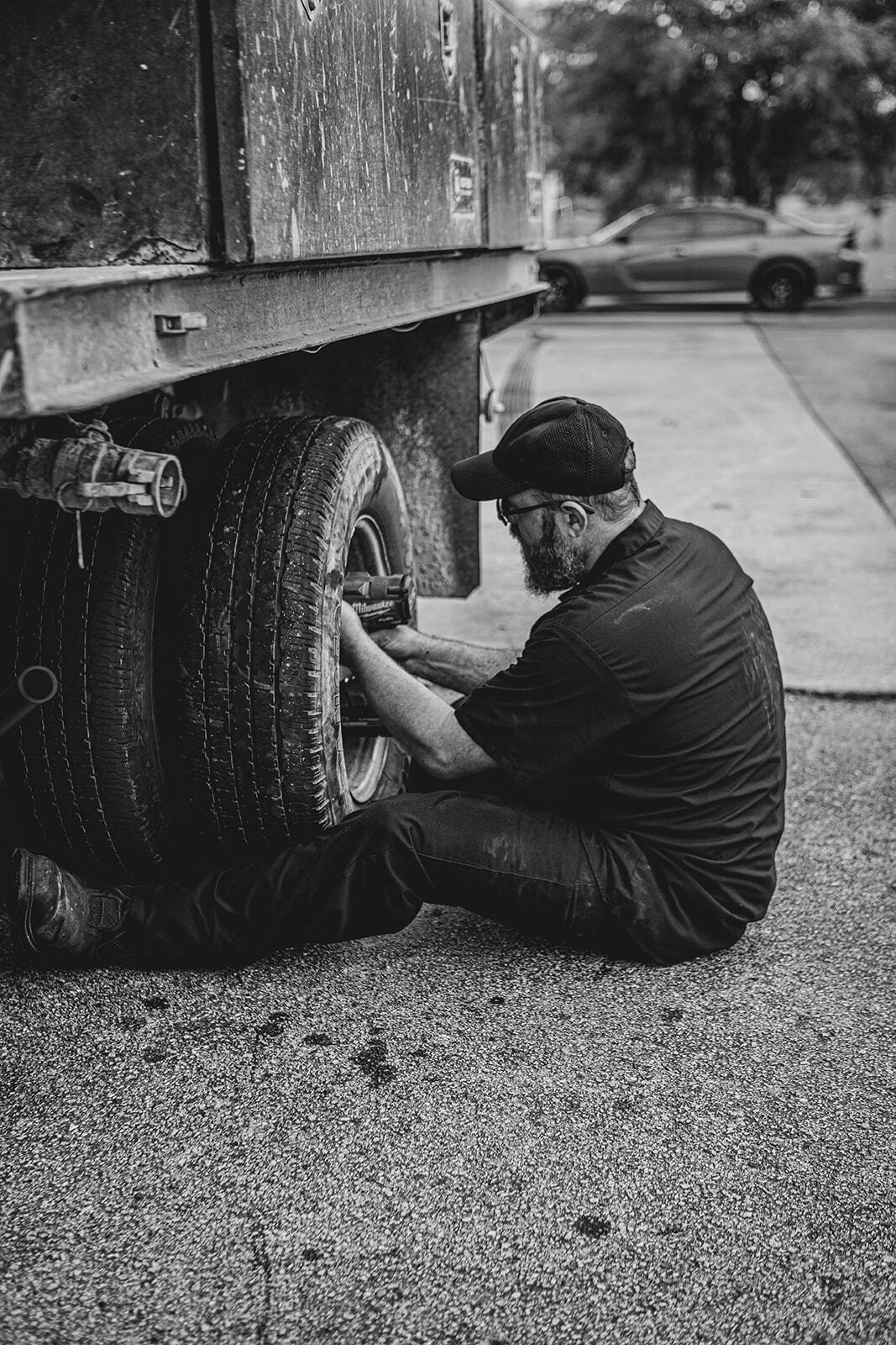 Man changing a truck tire on a gravel driveway. He wears a hat and dark clothing. A car is visible in the background.