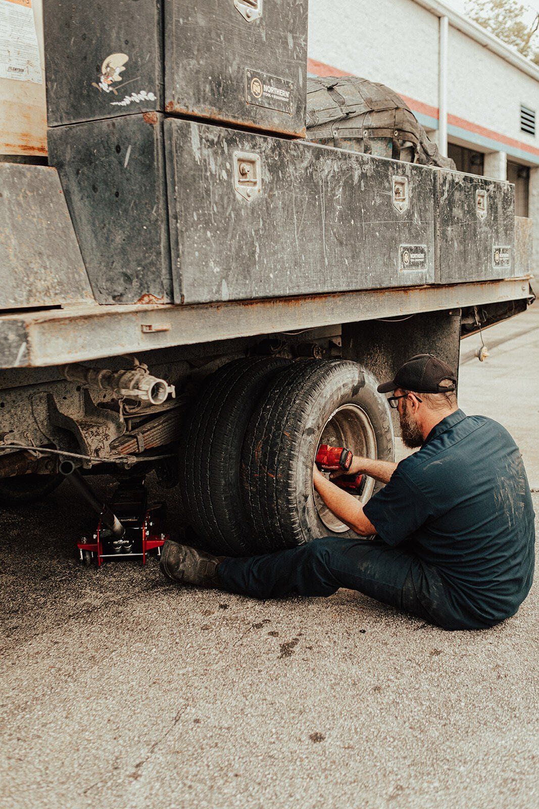 A person in a blue shirt changes a tire on a large, dirty vehicle, outside near a building.