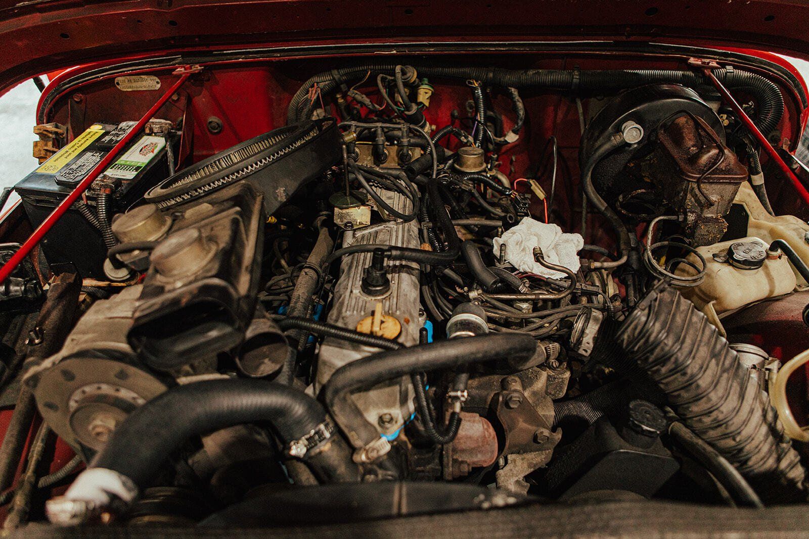 Red car engine bay filled with an engine, wires, hoses, and other mechanical parts.