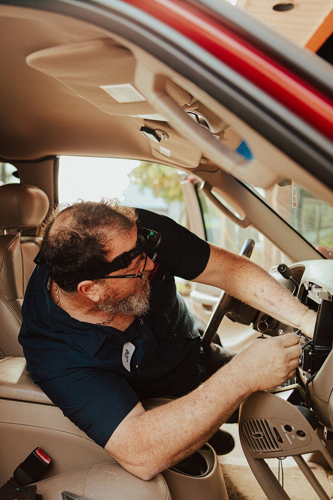 Man working on the dashboard of a red vehicle; wearing glasses and a blue shirt.