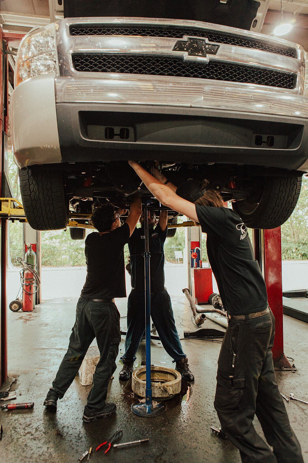 Three people work on the underside of a car lifted in a garage.