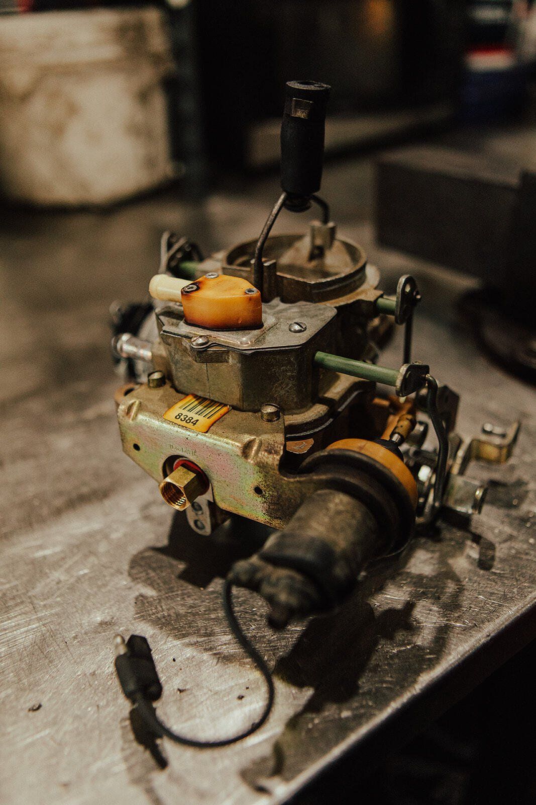 Carburetor on a metal surface, showing various components, wires, and attachments; mechanic's workshop background.