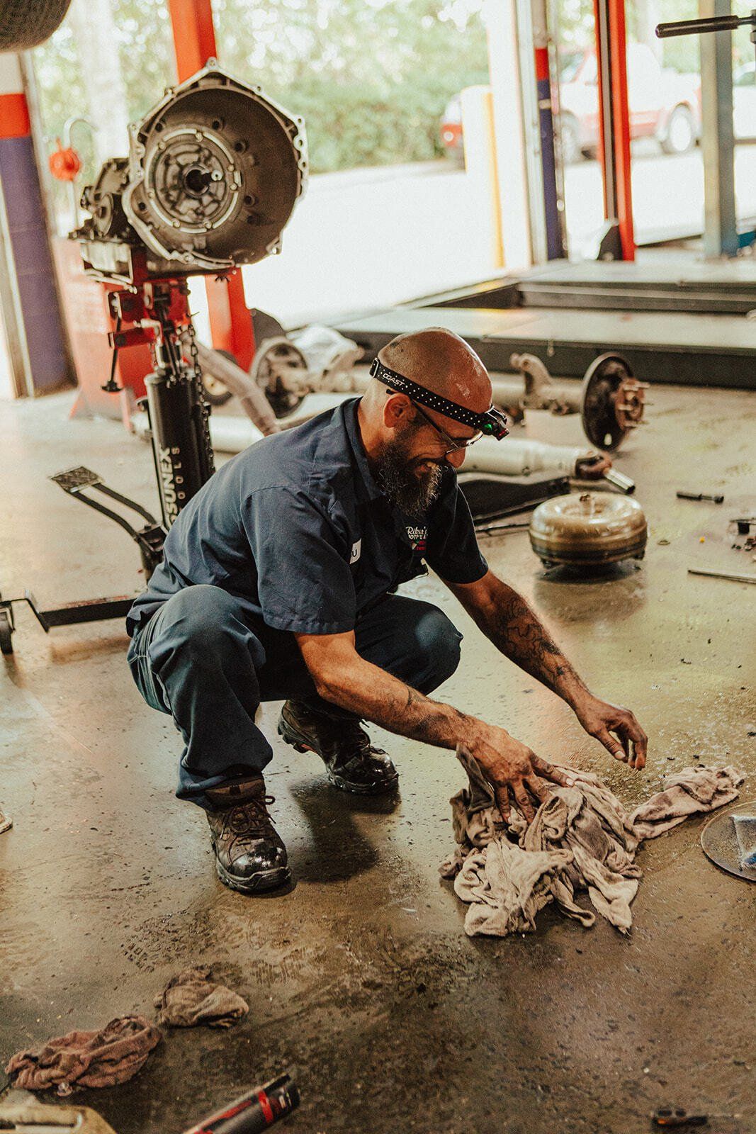 Mechanic in a garage, squatting and wiping a part with a cloth. Transmission parts and tools scattered around.
