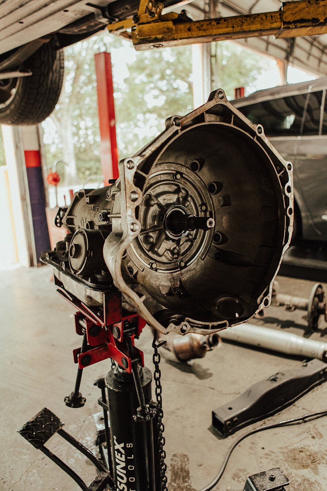 Transmission on a hydraulic jack in a garage, open and facing the camera, with tools and a car visible in the background.