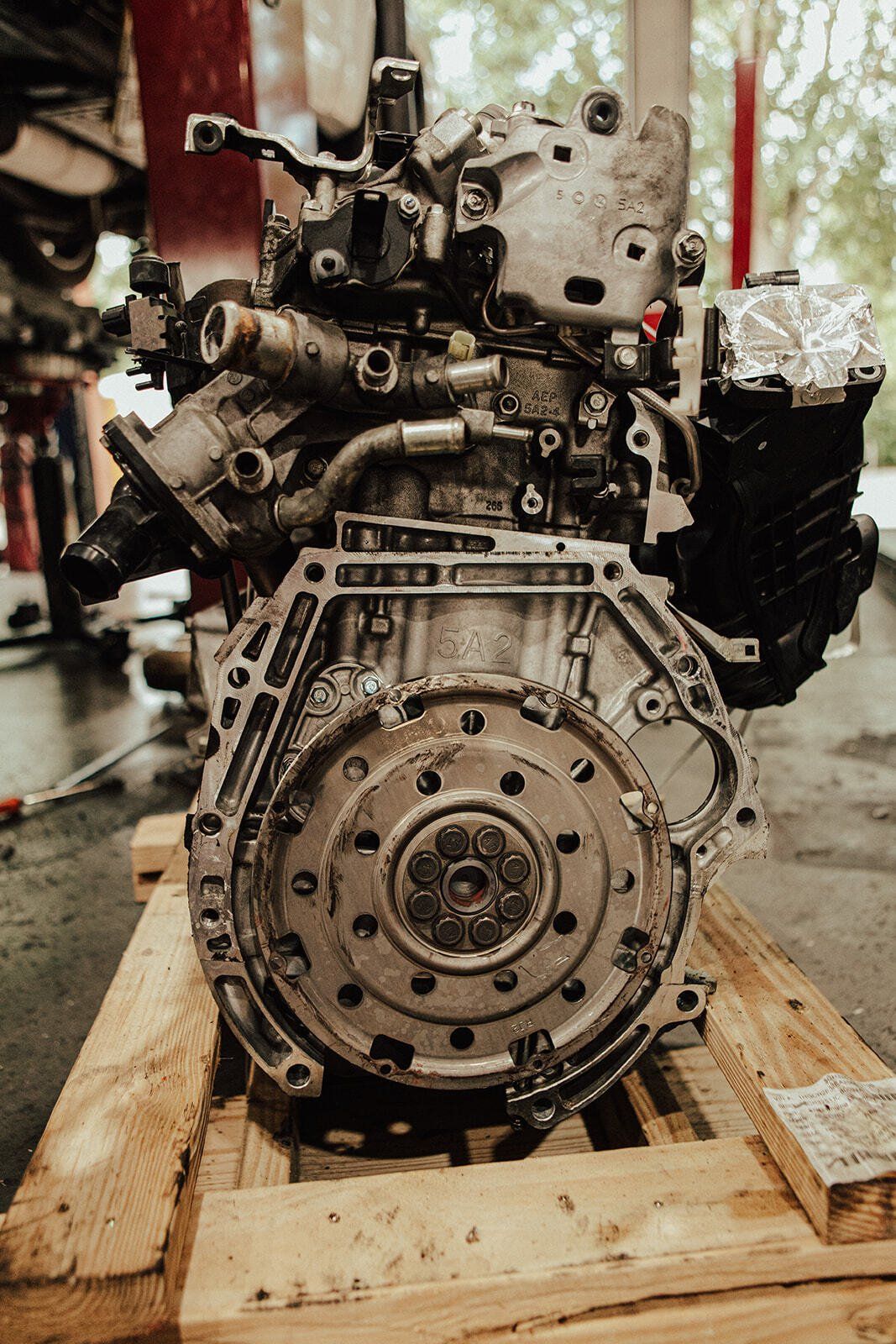 Car engine on a wooden pallet, viewed from the flywheel end, in an auto repair shop.