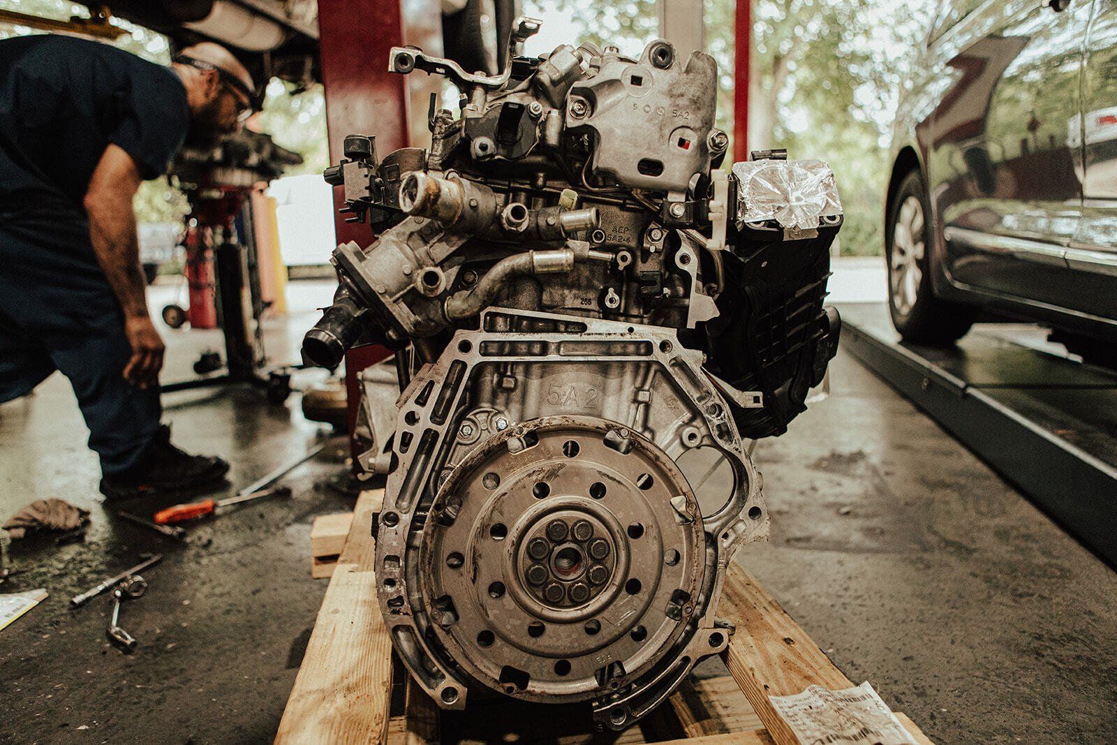 Car engine on wooden pallet in auto shop; mechanic working in the background.