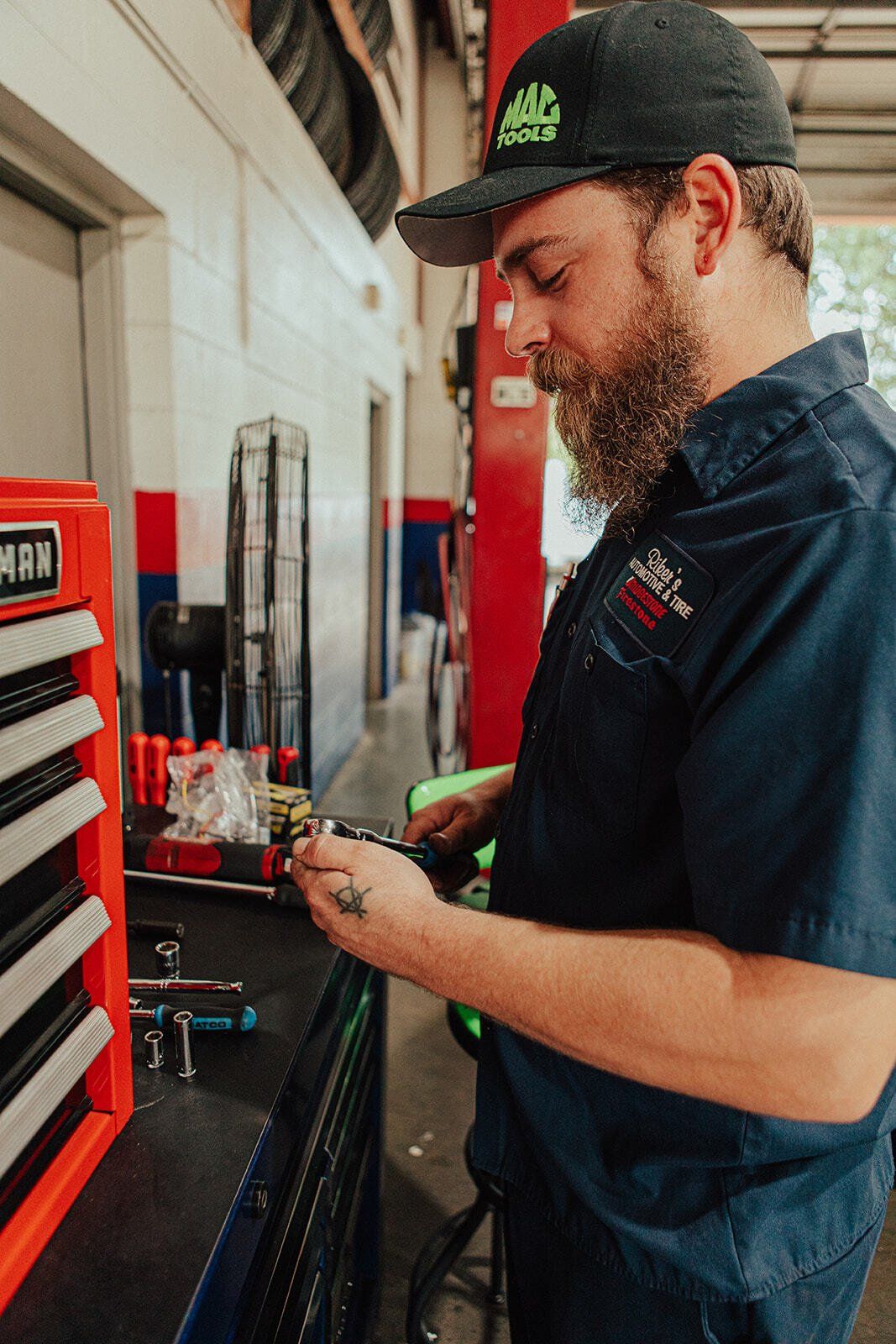 Mechanic in a shop examines a part, wearing a hat and work shirt. Red tool chest visible.