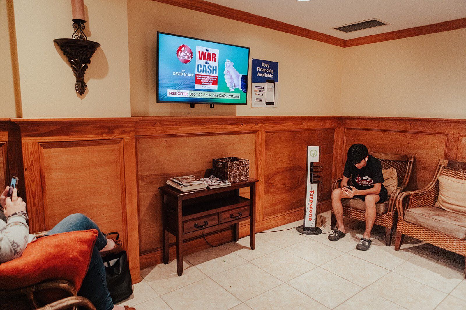 Waiting room with a person seated. A TV displays an advertisement, and there are magazines on a side table.