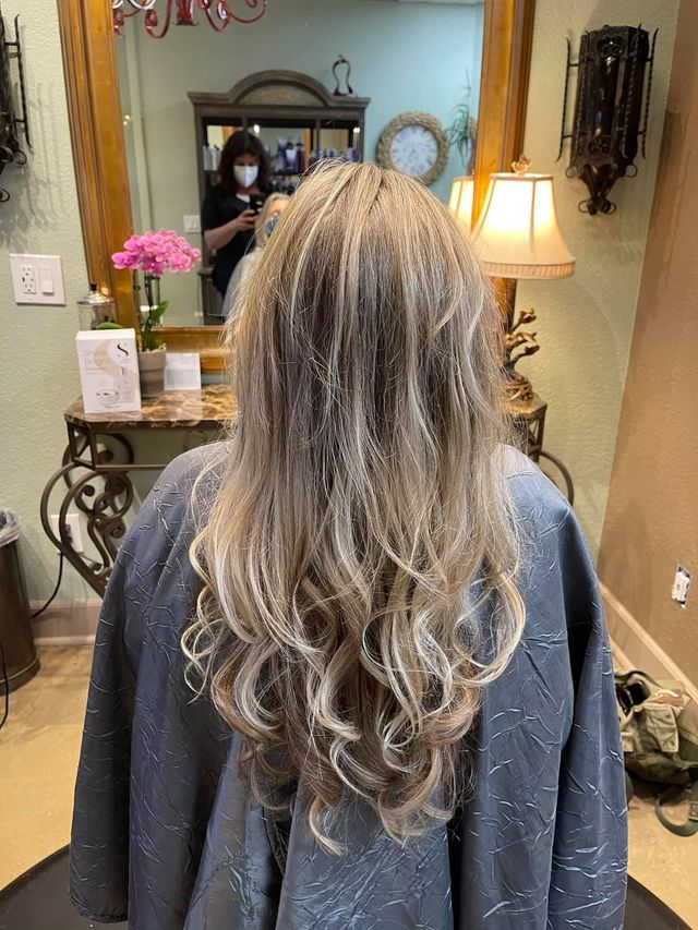 Woman with long, layered, highlighted hair at a salon. A stylist wearing a mask is reflected in the mirror.