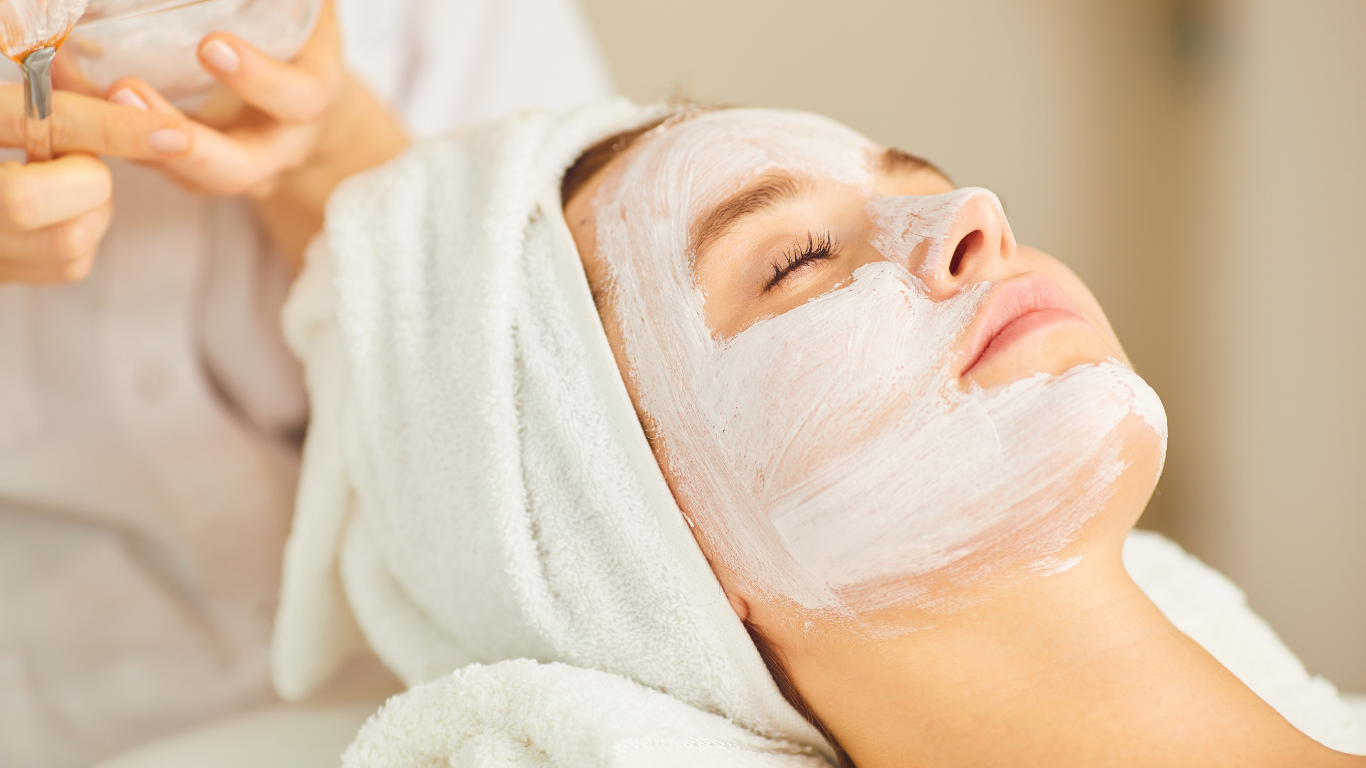 A person receiving a facial treatment at a spa; face covered in white mask, eyes closed, wrapped in a white towel.