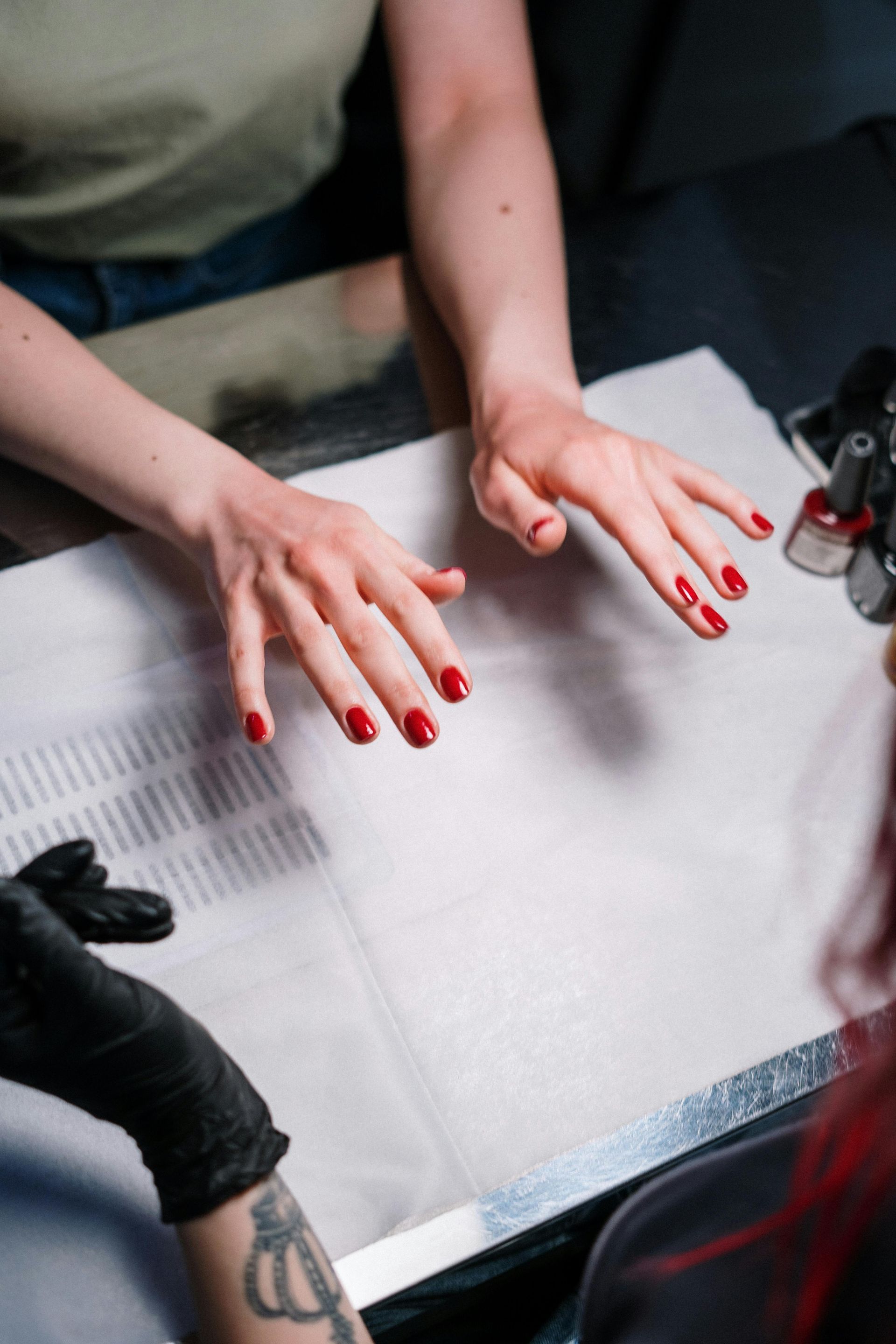 Person getting their nails painted red at a salon. Hands are centered, resting on a white surface, with a manicurist working.