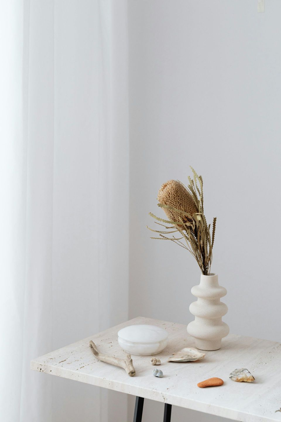 A beige vase with dried flowers, next to a white round container, rocks, and a wood piece on a stone table.