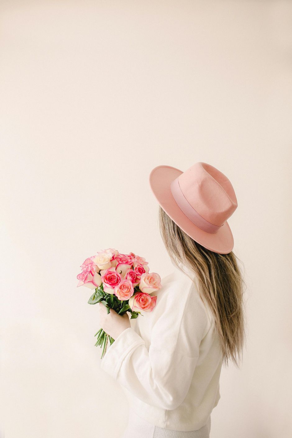 Woman in pink hat holding pink roses, standing against a cream background. Her back is to the camera.