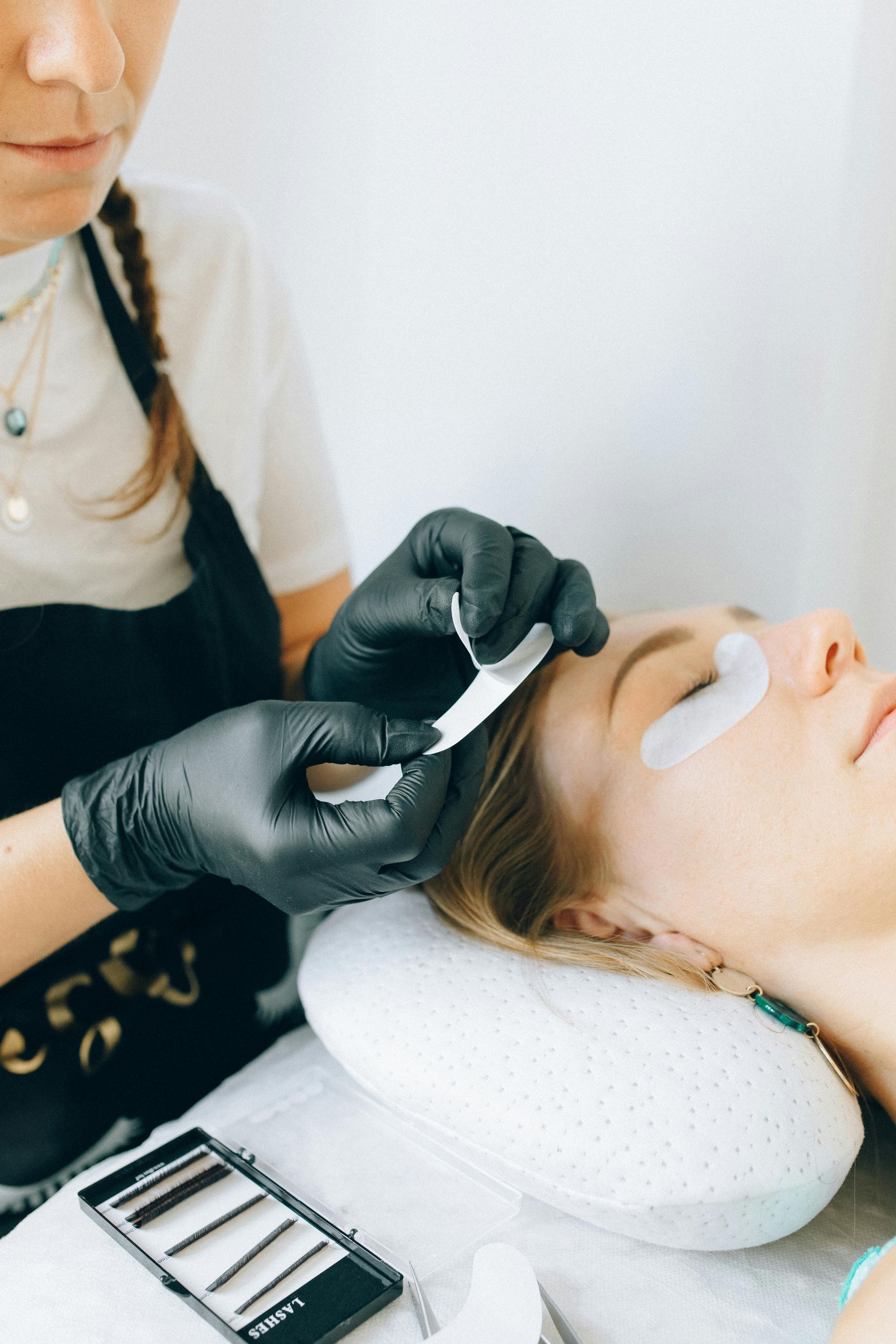 Woman receiving eyelash extensions; technician in black gloves applying a pad to the lower eyelid.