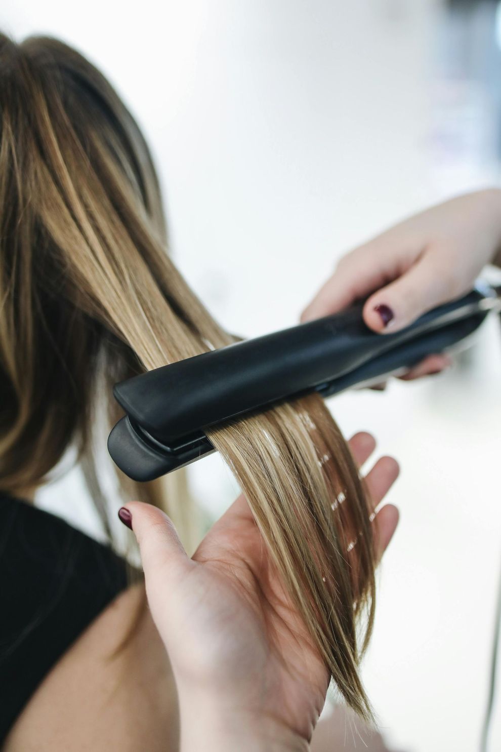 A person's hair being straightened with a black flat iron by a hand with painted nails.