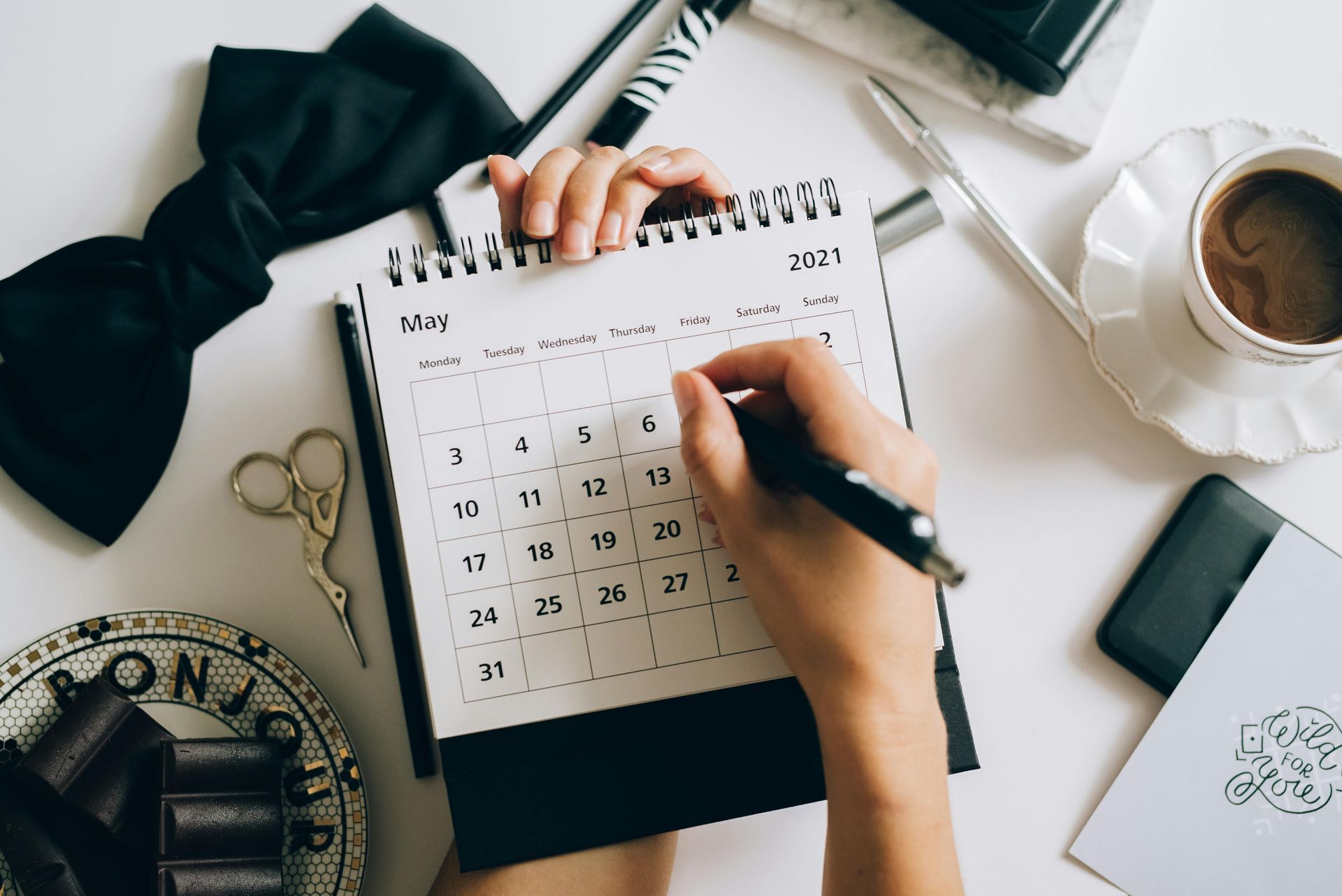 A person's hands writing on a May 2021 calendar on a white desk, with coffee, bow tie, and scissors visible.
